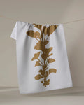 PrintedPlant’s “Oregano” is a white textured tea towel featuring a bold gold botanical silhouette, made from recycled materials and displayed hanging on a thin string against a neutral background.