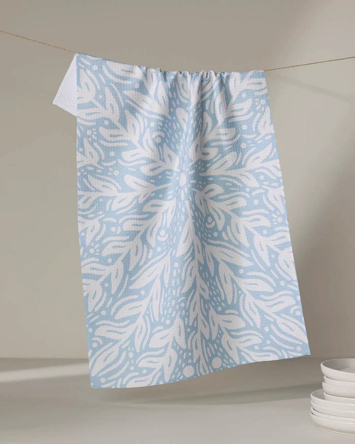The Outwards tea towel by Graphics and Grain, featuring a blue and white symmetrical leaf pattern, hangs from a line with white plates stacked nearby on a light surface.
