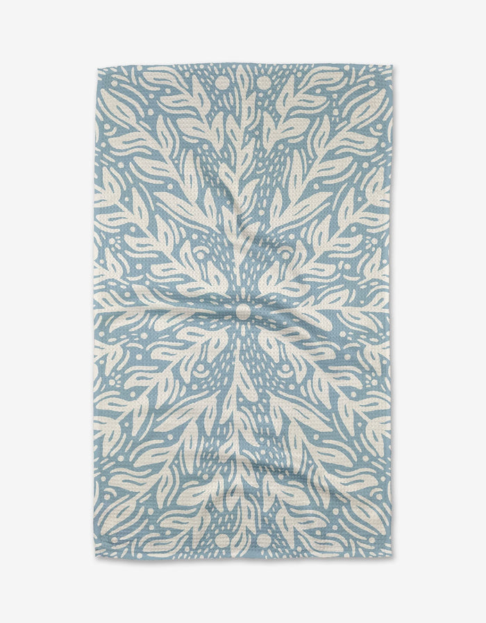 The Outwards tea towel by Graphics and Grain is rectangular, blue and white, and showcases a symmetrical botanical-inspired pattern with swirling leaves and dotted accents radiating from the center.