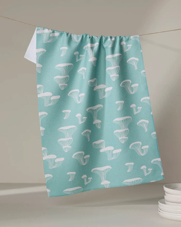 The Amberleigh Adoff Oyster Shroom tea towel, featuring a white mushroom pattern on light blue and made from recycled materials, hangs indoors above stacked white dishes against a neutral background.