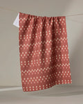 The "Paddles and Oars" tea towel by Coit Creative, made from recycled materials, features a reddish-brown hue with vertical cream dots and lines, and is shown hanging against a neutral gray background.