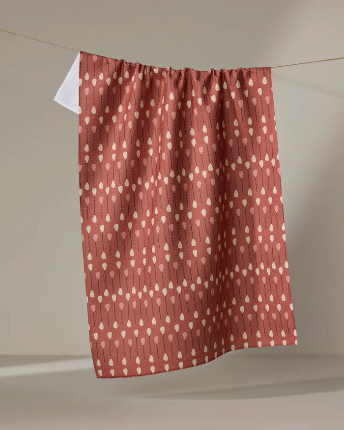 The "Paddles and Oars" tea towel by Coit Creative, made from recycled materials, features a reddish-brown hue with vertical cream dots and lines, and is shown hanging against a neutral gray background.