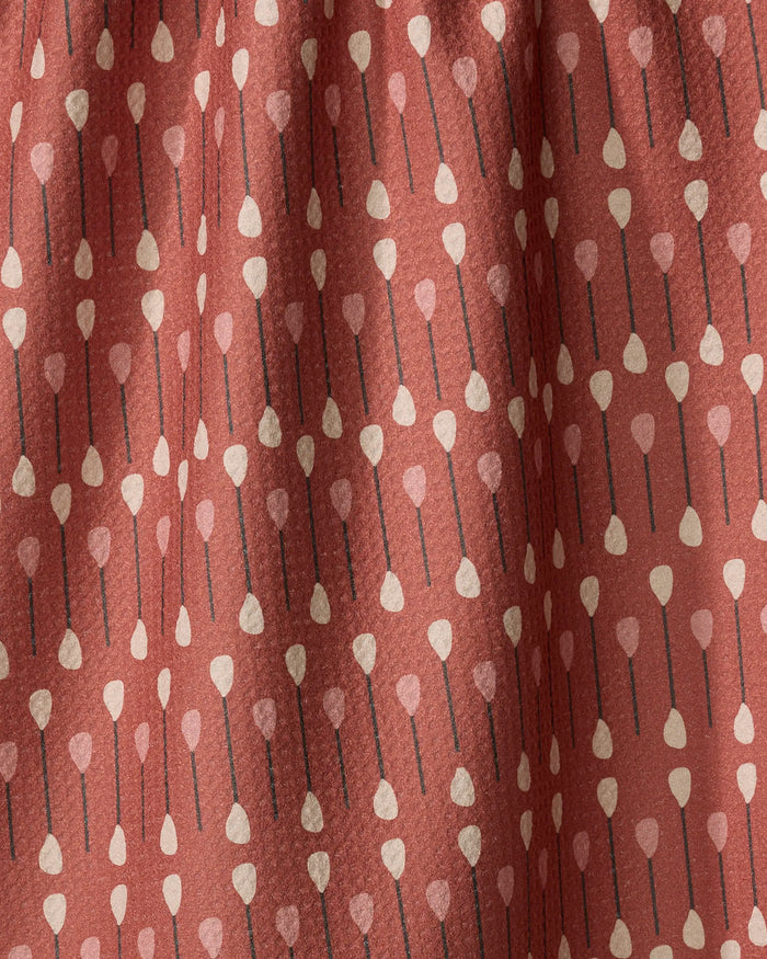 The Coit Creative "Paddles and Oars" tea towel features a rust color and vertical rows of stylized paddles with beige tips, black shafts, and light pink heart-shaped ends. Made from quick-drying recycled materials and shown with gentle folds.