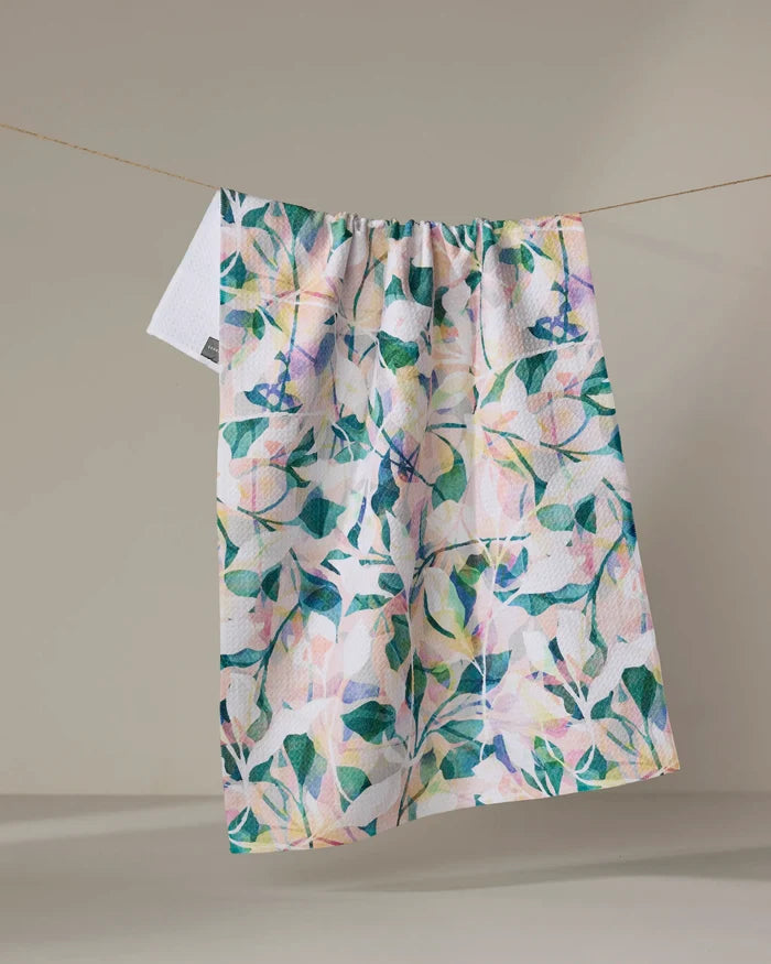 The "Painted Foliage" tea towel by Rachel Parker, crafted from recycled materials and featuring a leafy floral design, hangs on a clothesline against a neutral background.