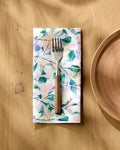 A fork with a wooden handle rests on Rachel Parker's Painted Foliage napkin, placed on a light wood table beside a beige ceramic plate. Sunlight casts soft shadows over the stylish Painted Foliage Dinner Napkin Set.