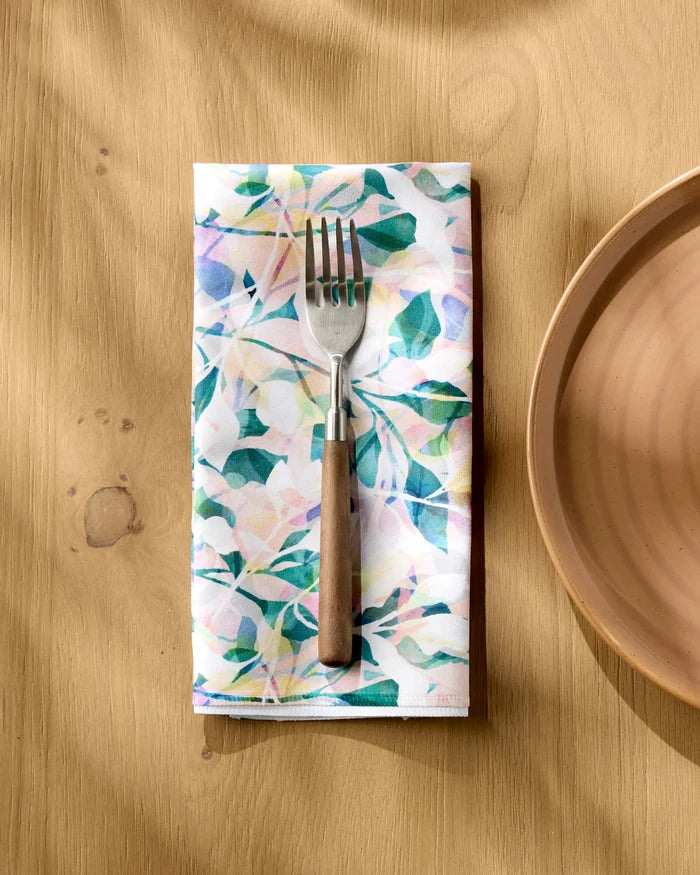 A fork with a wooden handle rests on Rachel Parker's Painted Foliage napkin, placed on a light wood table beside a beige ceramic plate. Sunlight casts soft shadows over the stylish Painted Foliage Dinner Napkin Set.