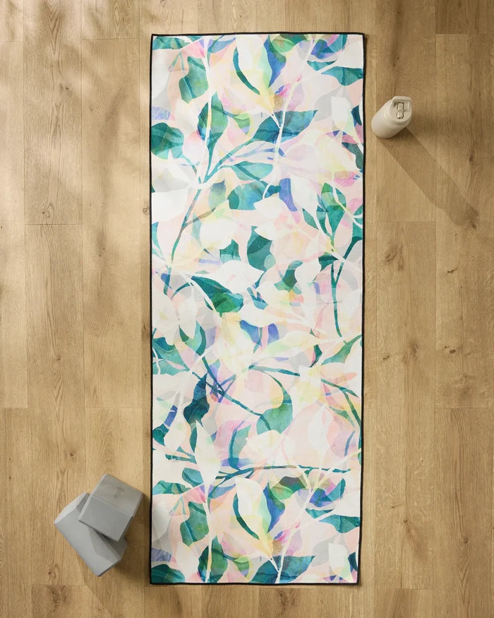 The Rachel Parker Painted Foliage yoga mat, featuring green, blue, and pink leaves on a light background, is spread on a wooden floor with a non-slip yoga towel, water bottle, and two gray blocks nearby for your practice.