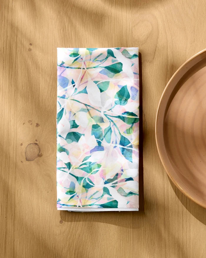 A neatly folded Painted Foliage recycled dinner napkin by Rachel Parker, featuring pastel floral patterns, rests beside a plain round beige plate on a light wooden table with sunlight casting soft shadows.