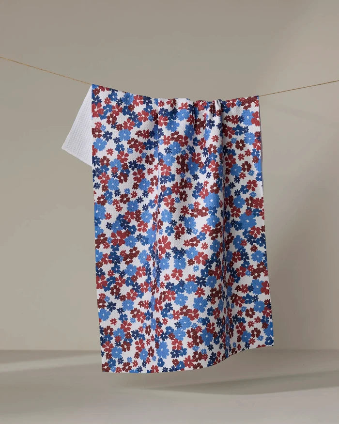 The Ashley Celena Painted Petals ultra-absorbent kitchen towel, featuring a small red, blue, and white floral pattern, hangs on a clothesline against a plain beige background.
