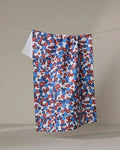 The Ashley Celena Painted Petals ultra-absorbent kitchen towel, featuring a small red, blue, and white floral pattern, hangs on a clothesline against a plain beige background.