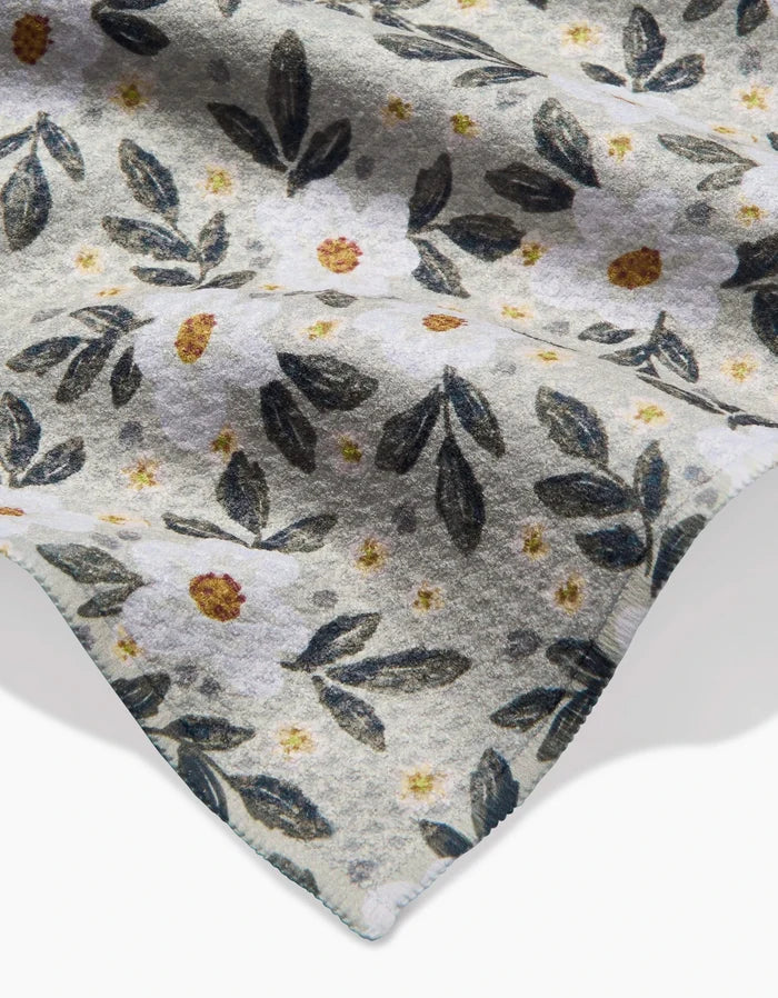 A close-up of Teri the Butter’s Painted Spring Flowers Geoweave bar towel, showcasing a soft fabric with white flowers, yellow centers, and dark green leaves on a light background. The corner edge appears at the bottom right.