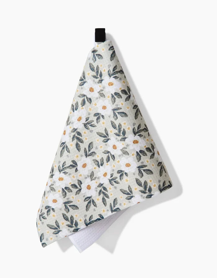 The Teri the Butter Painted Spring Flowers Bar Towel, featuring green leaves and white flowers on Geoweave fabric, hangs from a black loop against a white background.