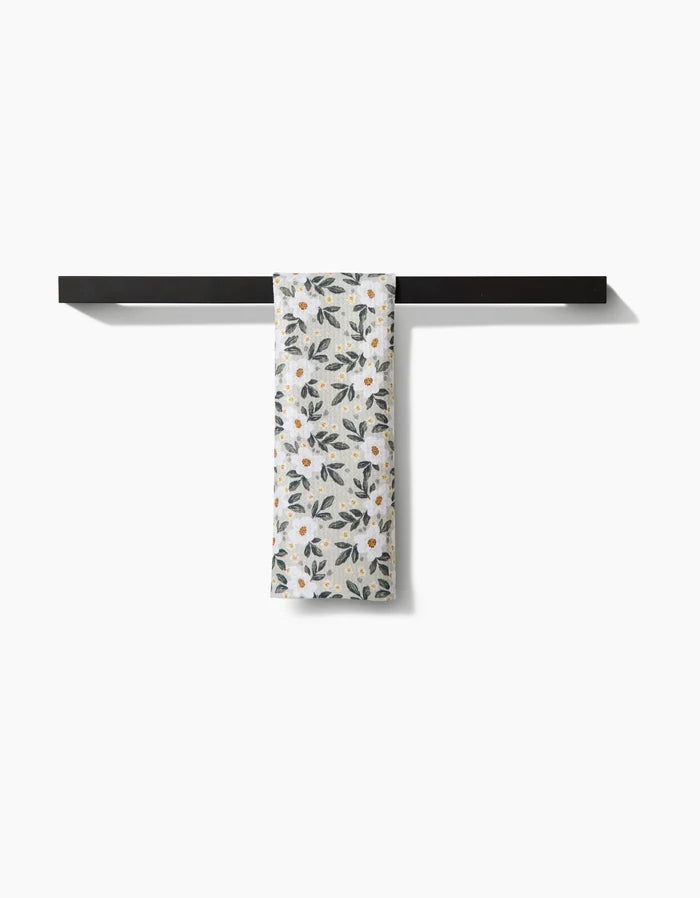 A white Painted Spring Flowers bar towel by Teri the Butter, adorned with blue and yellow blooms, is neatly draped over a sleek black towel bar against a plain white background.