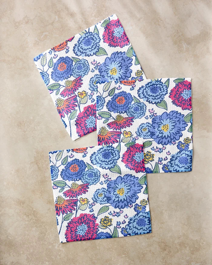 The Painterly Meadow napkins by Kris Ruff are made from recycled materials and feature a colorful floral pattern with blue, pink, and purple flowers and green leaves on a beige marble background.