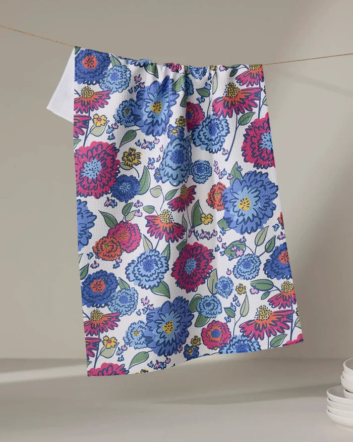 The Painterly Meadow tea towel by Kris Ruff features a vibrant pattern of blue, red, and pink flowers with green leaves, crafted from recycled materials. It hangs on a neutral background with stacked white dishes nearby.