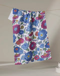 The Painterly Meadow tea towel by Kris Ruff features a vibrant pattern of blue, red, and pink flowers with green leaves, crafted from recycled materials. It hangs on a neutral background with stacked white dishes nearby.