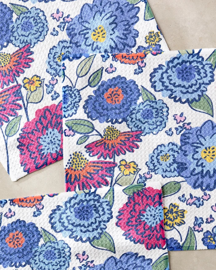 Three Painterly Meadow cloths by Kris Ruff, made from recycled materials and featuring a floral pattern of blue, purple, red, and yellow flowers with green leaves on a light beige surface, are overlapped at the corners.