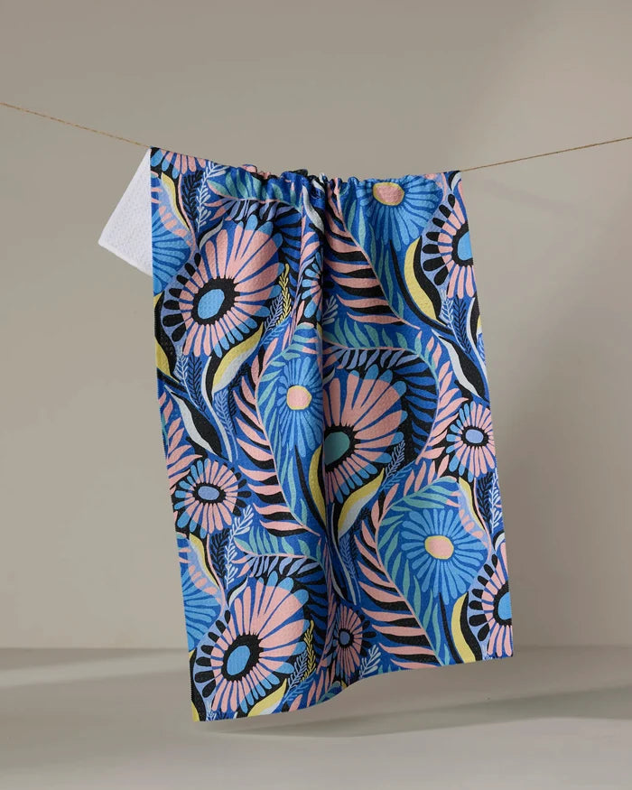 The Palm Beach kitchen towel by Masha Volnova, featuring a bold floral pattern in blue, pink, black, and yellow, hangs on a clothesline against a plain neutral background.