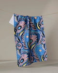 The Palm Beach kitchen towel by Masha Volnova, featuring a bold floral pattern in blue, pink, black, and yellow, hangs on a clothesline against a plain neutral background.