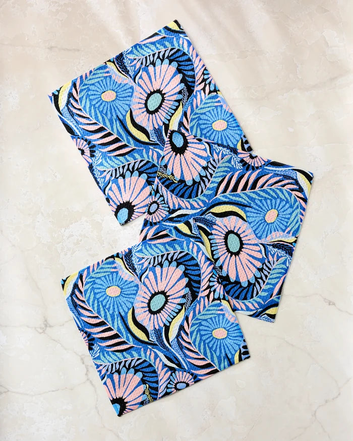 Three "Palm Beach" fabric napkins by Masha Volnova, featuring a vibrant floral pattern in blue, pink, yellow, and black, are displayed on a light marble surface—perfect for brightening any table with bold color.