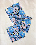 Three "Palm Beach" fabric napkins by Masha Volnova, featuring a vibrant floral pattern in blue, pink, yellow, and black, are displayed on a light marble surface—perfect for brightening any table with bold color.