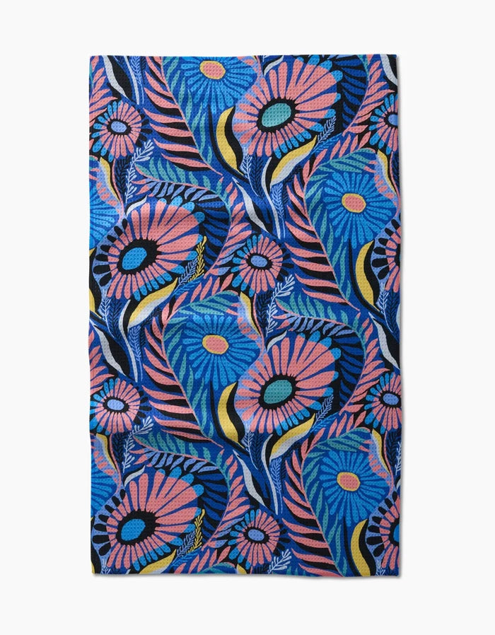 The Palm Beach tea towel by Masha Volnova features a vibrant floral pattern with large blue, pink, and yellow flowers and green leaves on a blue background, crafted from recycled materials.