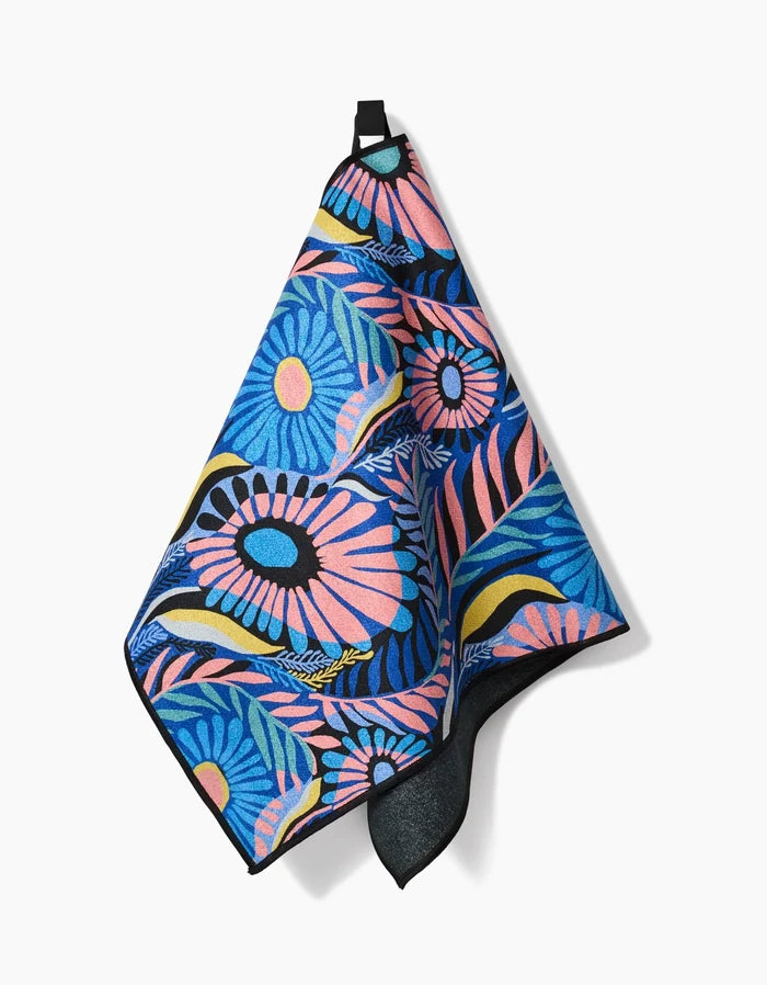 The Palm Beach towel by Masha Volnova features a bold blue, pink, yellow, and black floral pattern and hangs from a black loop—colorful and ultra-absorbent, it’s perfect as a fitness towel.