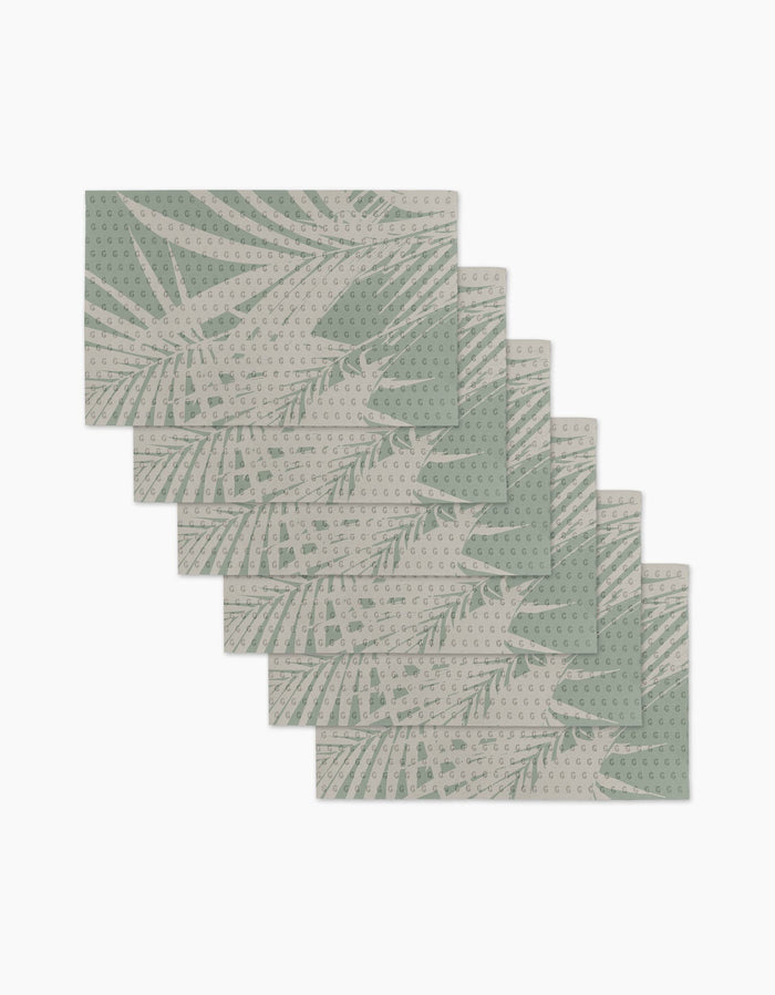 Six Palm Breeze rectangular placemats by Geometry are stacked in a fan arrangement, featuring a light green botanical leaf pattern on beige with dotted details. Made from recyclable materials, they offer an eco-friendly alternative to paper products.