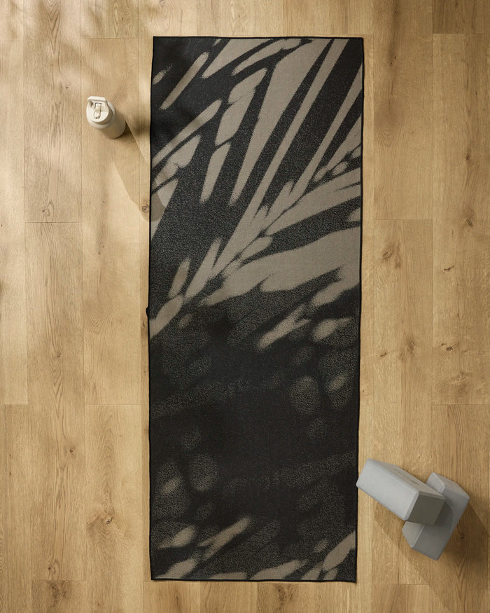The GEOMETRY Palm Shadow yoga mat in black and gray is laid out on a light wood floor, with a non-slip yoga towel, gray yoga block, and light-colored water bottle placed nearby.