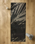 The GEOMETRY Palm Shadow yoga mat in black and gray is laid out on a light wood floor, with a non-slip yoga towel, gray yoga block, and light-colored water bottle placed nearby.