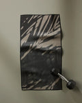 The GEOMETRY Palm Shadow ultra-absorbent fitness towel in a black and beige pattern is laid flat on a gray surface, with a black hexagonal dumbbell resting on its lower right corner.