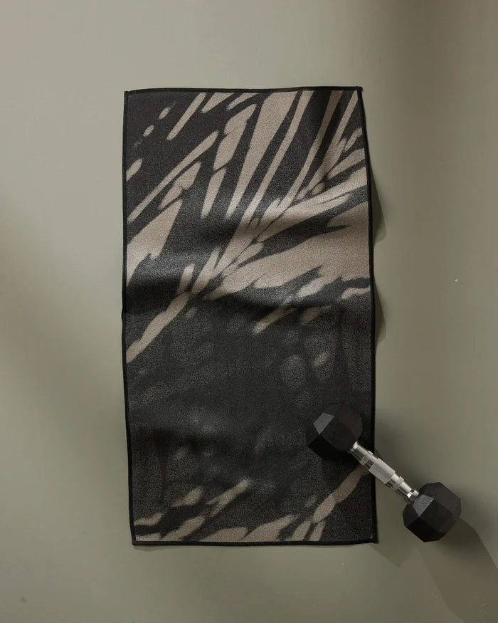 The GEOMETRY Palm Shadow ultra-absorbent fitness towel in a black and beige pattern is laid flat on a gray surface, with a black hexagonal dumbbell resting on its lower right corner.