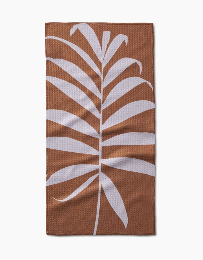 The PrintedPlant Palm bar towel is a brown rectangular towel made from ultra-absorbent Geoweave fabric, featuring a large white palm leaf design in the center and shown on a white background.