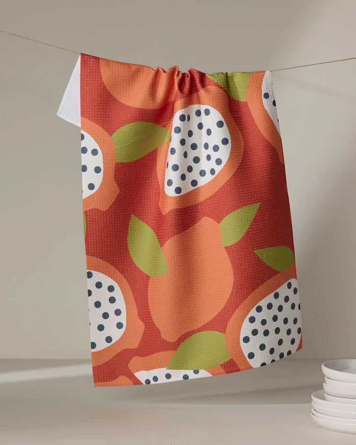 The Lizzie Clark Papaya Playtime kitchen towel features a vibrant orange, green, and white fruit pattern with dotted accents. It hangs on a string, while stacked white plates appear in the lower right corner.