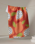 The Lizzie Clark Papaya Playtime kitchen towel features a vibrant orange, green, and white fruit pattern with dotted accents. It hangs on a string, while stacked white plates appear in the lower right corner.