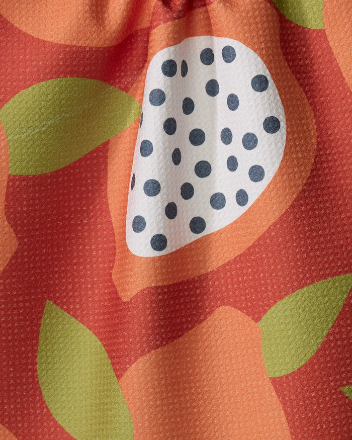 A close-up of the Lizzie Clark "Papaya Playtime" tea towel shows its absorbent texture, colorful abstract fruit shapes, green leaves, and a white oval with black dots on a red background. Visible folds highlight its quality design.