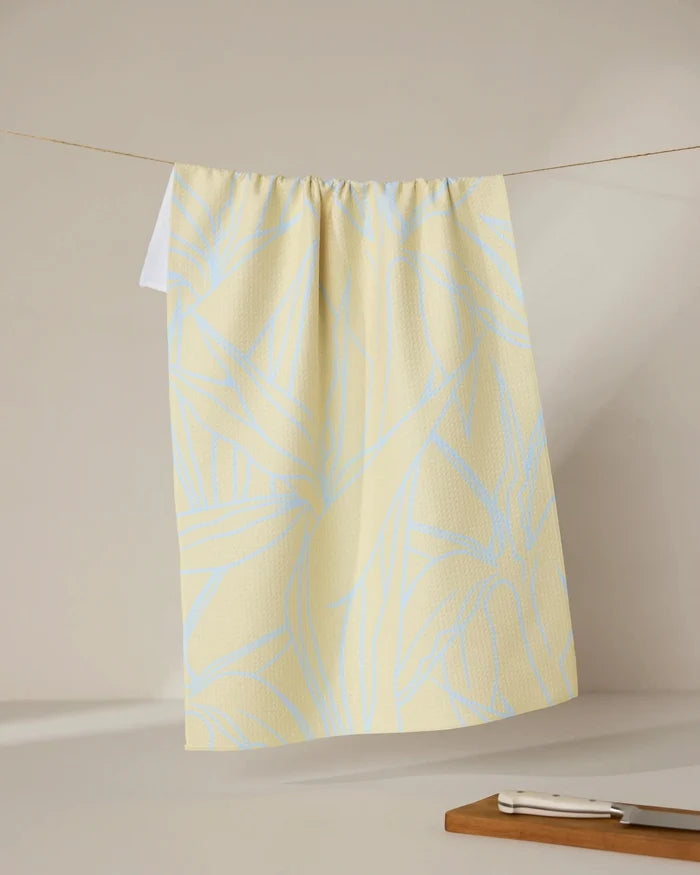 The GEOMETRY Paradise Blooms tea towel, featuring a light blue abstract leaf pattern on yellow, hangs on a line with a cutting board and knife placed nearby against a neutral background.