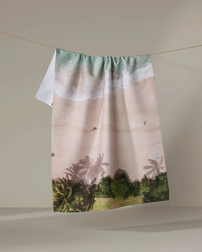 The Kait Hanson "Paradise Found" towel, featuring ocean waves, a sandy shore, and palm trees, dries quickly and hangs on a line against a plain background.