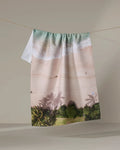 The Kait Hanson "Paradise Found" towel, featuring ocean waves, a sandy shore, and palm trees, dries quickly and hangs on a line against a plain background.