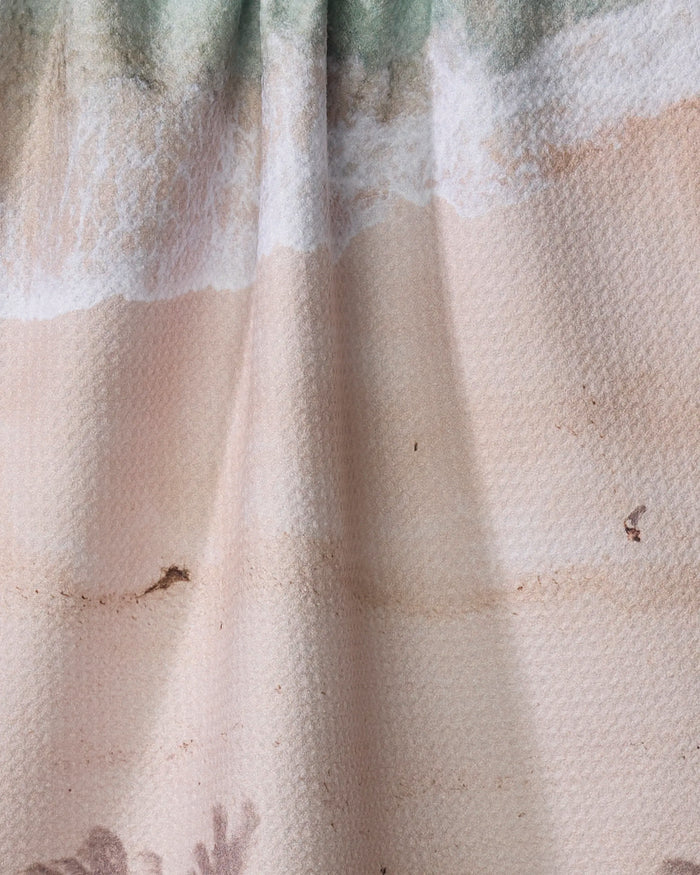 A close-up of the Paradise Found quick-drying towel by Kait Hanson, featuring a beach scene with waves, sand, and footprints. Its textured waffle-like surface slightly distorts the print due to fabric folds.