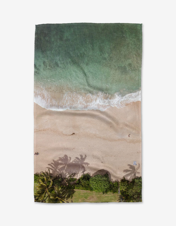 Aerial view of Kait Hanson’s Paradise Found beach towel, made from post-consumer recycled materials and featuring a printed tropical coastline scene with waves, sand, and palm tree shadows.