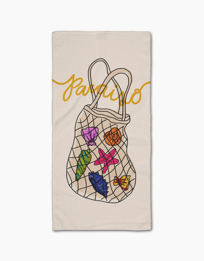The Geometry Paraiso beige bar towel, made from post-consumer recycled materials, features a mesh bag with colorful seashells and leaves, and the word paraiso in yellow cursive above.