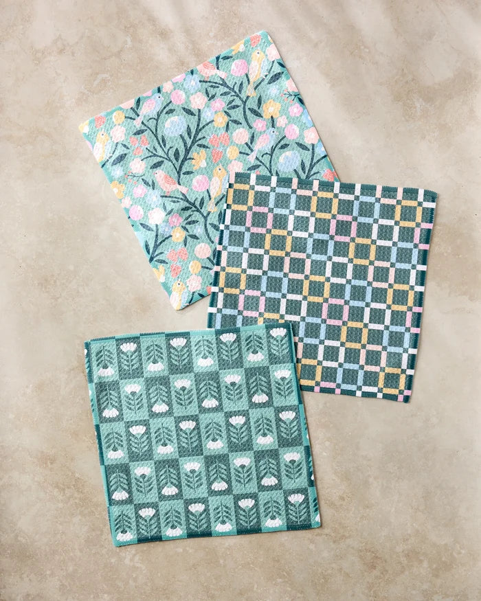 Three patterned Park Lane dishcloths by Charlie Rowan Designs, made from recycled materials, are laid on a beige surface: one with pink and yellow florals, one checkered, and one with white tulips on green.