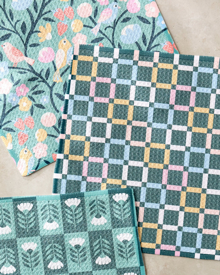 The Park Lane dishcloth set by Charlie Rowan Designs features three patterned cloths—birds and flowers, pastel plaid, and white flowers on green—all made from recycled materials and shown slightly overlapping on a neutral surface.
