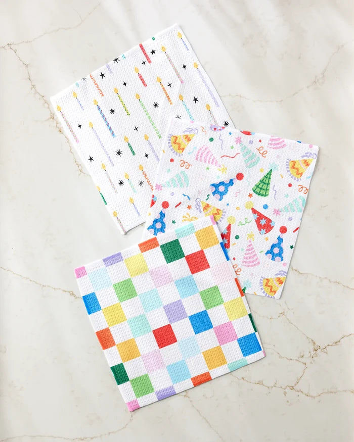 Three colorful Party Pop cloth napkins by Vaishali Design Studio, made from recycled materials, are arranged on marble—one with a multicolored grid, one party-themed, and one with small colorful lines and crosses.