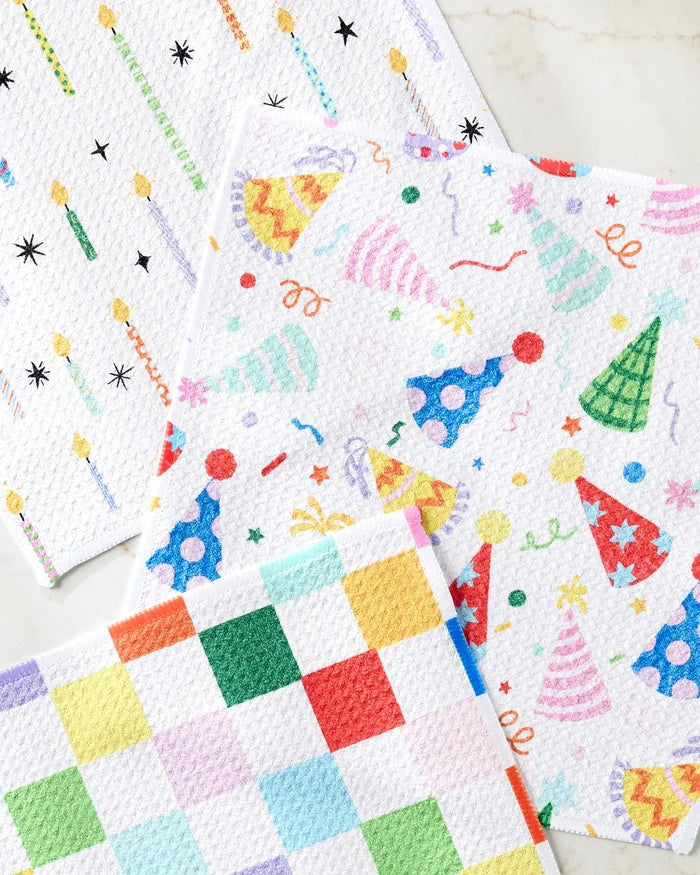 Three colorful Party Pop towels from Vaishali Design Studio, made from recycled materials, are laid out on a white surface—one with candles, one with party hats and confetti, and one with a multicolored checkered design.