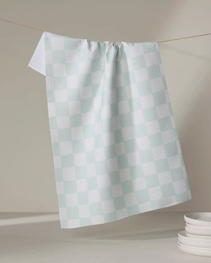 The Sweet Simple Vegan Pastel Picnic tea towel in light blue and white checkered pattern hangs on a string, with a stack of white plates nearby on a neutral surface.