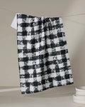The Patch Ash recycled kitchen towel by Geometry in a black and white checkered pattern hangs on a string against a neutral background, with white dishes stacked in the lower right corner.