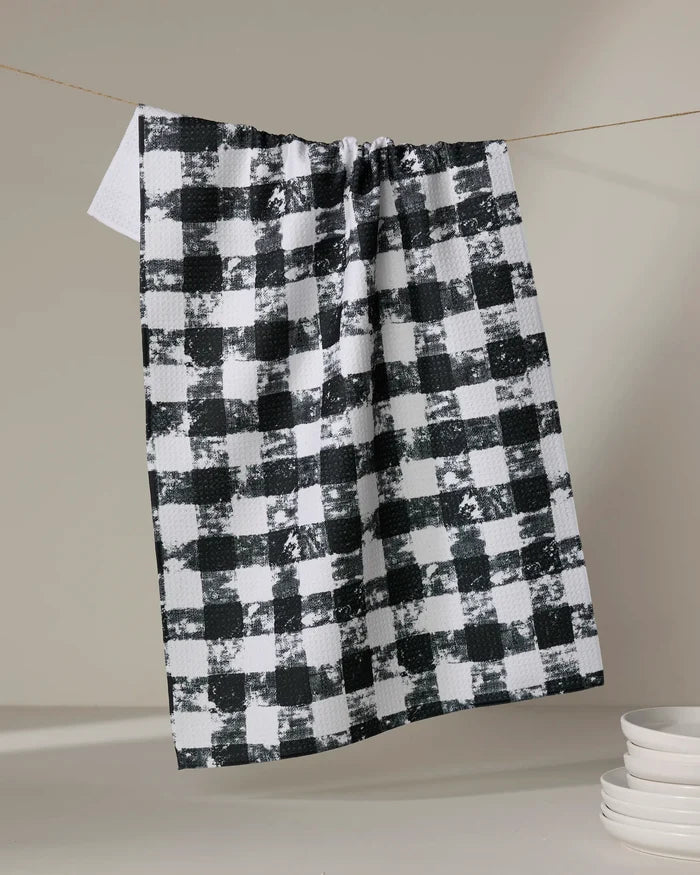 The Patch Ash recycled kitchen towel by Geometry in a black and white checkered pattern hangs on a string against a neutral background, with white dishes stacked in the lower right corner.