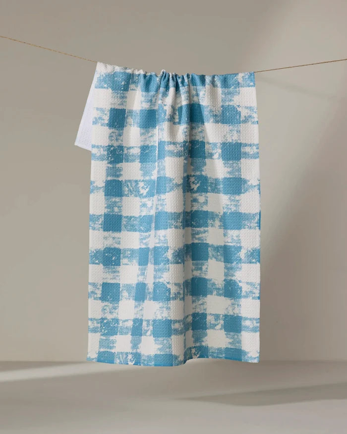 The Geometry Patch Denim recycled kitchen towel, featuring a blue and white checkered textured pattern, hangs on a clothesline against a neutral background.